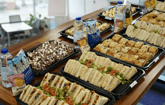 A spread of sandwich platters with a selection of fresh snacks, bottled drinks, and crisps, ideal for office events.